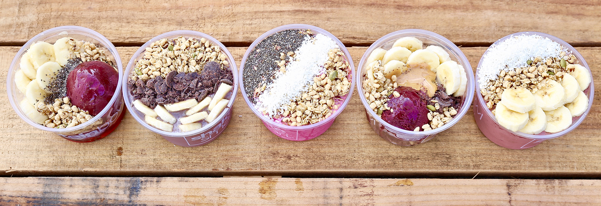 A variety of Shake Smart acai bowls displayed in a line on top of a wooden table